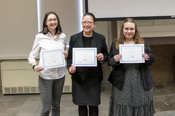 Three students stand in front of a fire place with certificates