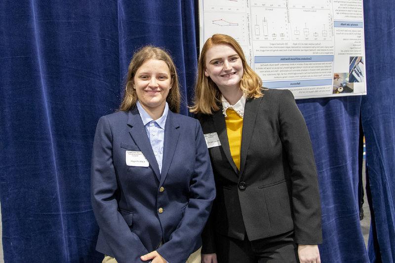 Two students wearing suits pose for a photo in front of a research poster.