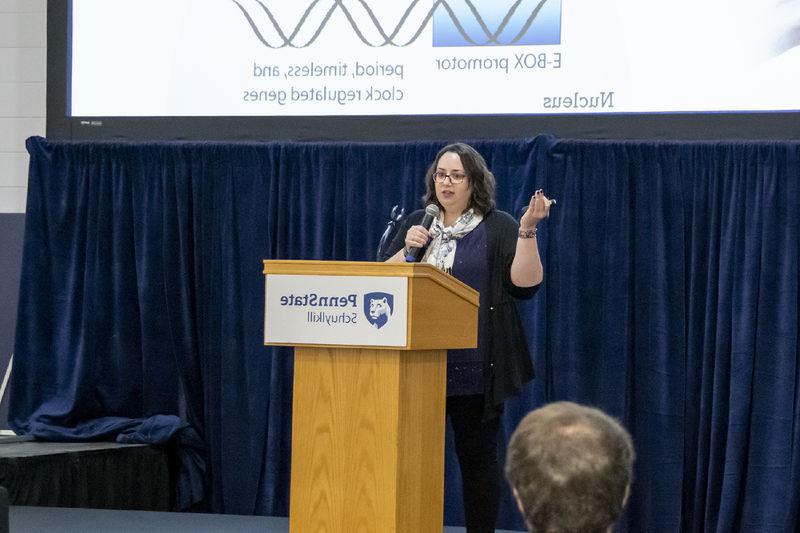 Woman stands in front of a projector screen and behind a podium, 拿着麦克风, 她指着她的ppt幻灯片